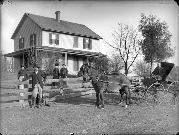 Man posing sitting in a buggy pulled by a single horse. Nearby are two men and two women posing standing near a wooden fence. In the background is a two-story frame house.