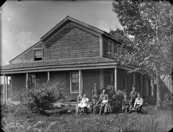 Group portrait of man, a woman holding a child, and two boys posing sitting, and behind them a girl and boy posing standing. They are in the yard in front of a frame house.	
