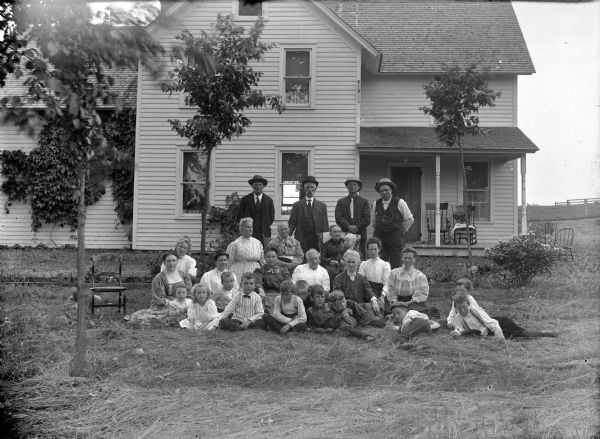 Group portrait of men, women and children posing sitting and standing in the yard in front of a two-story frame house. Probably a family reunion group.	