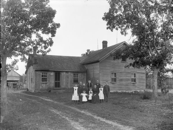 Man and woman posing sitting, surrounded by six girls and three boys in the yard in front of a board and frame house.
