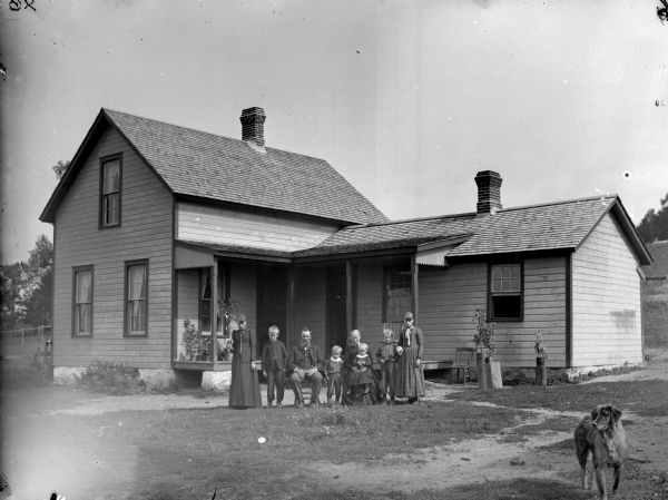 A man, and a woman holding a child, are posing sitting. They are surrounded by two girls and three boys in the yard in front of a house. There is a dog standing in the right foreground.
