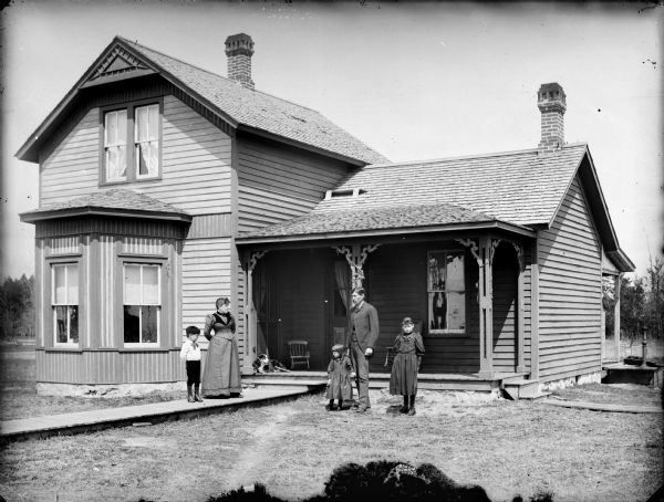 A woman and young boy are standing on a wooden walkway, and a man is standing in the yard to the right with two young girls. A dog is lying on the porch of the house behind them. On the right along the side of the house is a water pump on a platform near a rear porch.