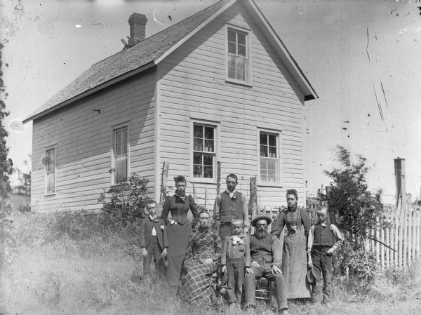 Man and woman posing sitting. They are surrounded by two women, three boys, and a man in a yard in front of a frame building.	