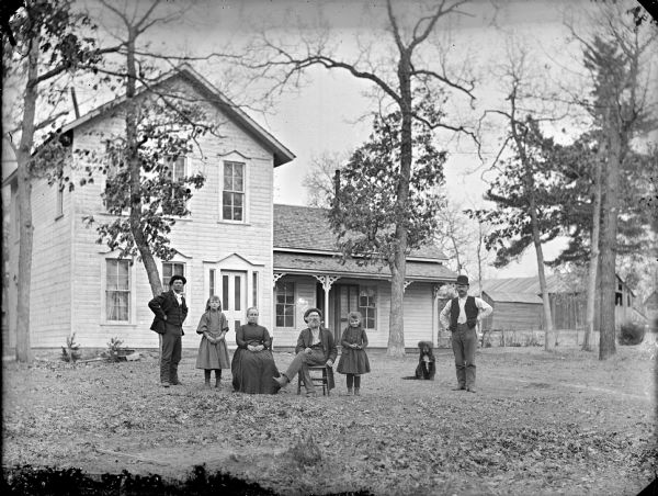 Man and woman posing sitting, and two men and two young girls standing, along with a dog in a yard in front of a frame house with a porch. Farm buildings are in the background.