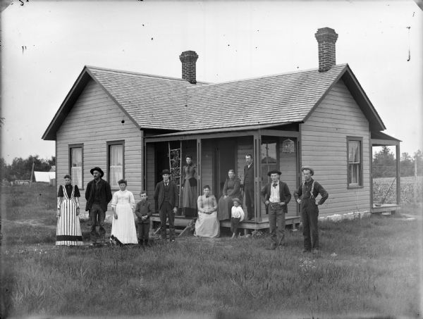 Two women are posing sitting, and three women, five men, and two young boys are posing standing on and near the porch of a small frame house. On the right is the side of another porch on the opposite side of the house. In front of one of the women standing there appears to be a dog playing with a stick. A bird is in a birdcage hanging from the porch.
