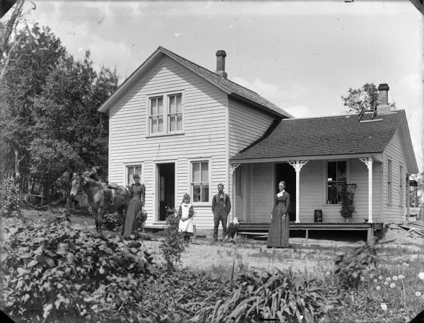 Two women, a man and young girl are posing standing in the yard in front of a frame house with a small porch. The woman standing on the left is holding a hat and is standing next to a horse with a saddle and flowers in its bridle. In the center on the ground a stuffed/mounted eagle is displayed.