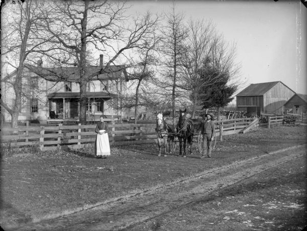 Man and woman posing in front of a fence. They are standing near a wagon pulled by a team of two horses. In the background is a two-story frame house with a porch, a large wood pile, a covered carriage, and farm buildings.