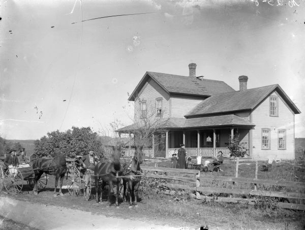 On the far left a man and woman are posing sitting in a buggy pulled by a single horse. Next to them is a man posing sitting in a buggy pulled by a team of horses. They are in front of a wooden fence enclosing a yard, where a man is posing sitting and reading a newspaper. Nearby a woman and girl standing next to three empty chairs. In the background is a two-story frame house, with a bird in a birdcage hanging from the porch roof. Negative marked with ink "No. 25-8."