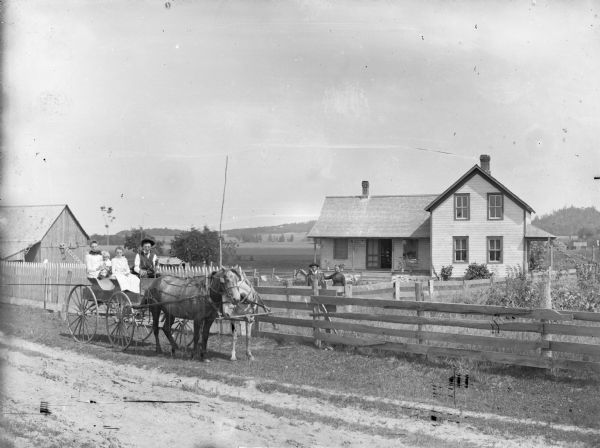Two young girls, a man and an infant are posing sitting in a wagon pulled by a team of two horses wearing fly-nets. On the right a man and a woman are posing standing behind a fence in the yard in front of a frame house. There is a baby carriage on the porch. In the far background are fields and hills.