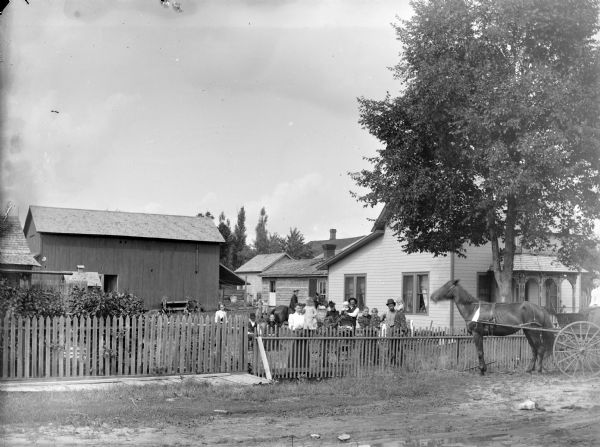 View of a group of people posing behind a fence. In the background is a frame house and farm buildings. There is a woman and a man posing standing, each holding a child, and a man and woman posing sitting. There are two women standing, and a man on horseback. A horse pulling a buggy is standing in front of the fence on the right.