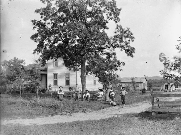 View across dirt road or path towards two men and a child who are posing sitting, with a woman, girl, and boy standing near a baby in a baby carriage. The boy is holding a rope tied to a cow that is drinking from a bucket. The group is under a large tree in front of a two-story frame house, with farm buildings and a windmill in the background.	