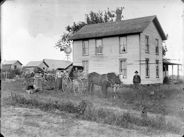 A man, woman, and two children are posing sitting in a buggy pulled by a team of two horses. A man is standing near the corner of the two-story frame house, and two women with a dog are standing on the left. In the background there is a windmill on the roof of a barn, other farm buildings, and a large pile of fuelwood.