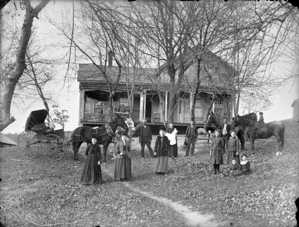 Family posing in yard of house with four horses, with two pulling a horse-drawn carriage. There are four women, two men, a young man, five young girls, and an infant. Three of the women are holding their gloves in their hands, and the other woman is holding the infant in her arms. One young girl is sitting bareback astride a horse on the far right. The house has a porch and is up on a small rise above the yard. There is a pedal grinding wheel in the yard.