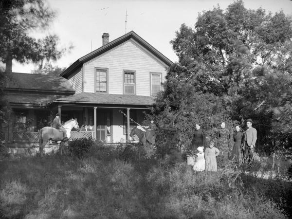 Boy and woman posing sitting on separate horses, and three women, two girls, and a man posing standing. They are in front of a frame house, and a man is posing sitting on the porch.