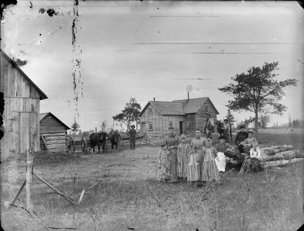 A farm family posing in front of farm buildings and farmhouse. The young daughters are wearing matching dresses. Men in the background are standing with a team of oxen pulling a wagon, and in the background on the right two people are standing with two horses.