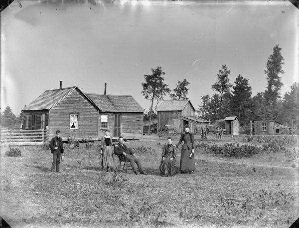 Man and woman posing sitting, and a woman, boy, and girl posing standing. In the background is a frame house and farm buildings.