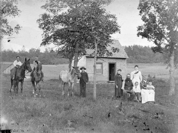 Two young boys are posing sitting on two horses, and a third boy is standing and displaying a horse. On the right a man and woman are posing sitting, with the woman holding an infant in her lap. Beside them are standing two girls and a young boy.