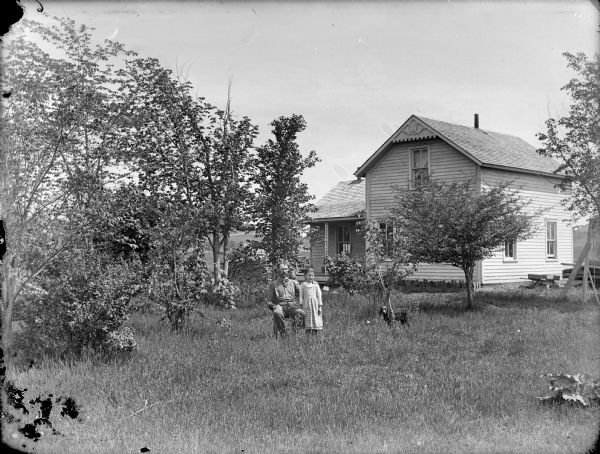 View across yard towards a man posing sitting, and a girl and dog standing. In the background is a frame house.