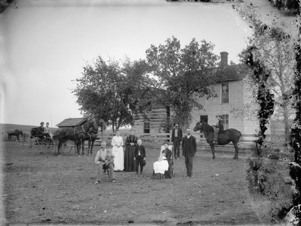 In the foreground a man and woman are posing sitting. They are each holding a young child. Behind them posing standing are two women, two men, and a boy. They are flanked by a boy on horseback, and a man and woman in a buggy pulled by a team of two horses. In the background is a wooden fence and two-story frame house.