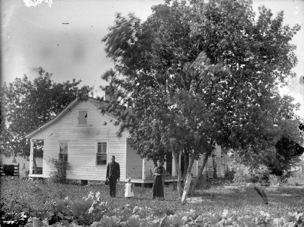 Man, woman, and small child posing standing in a yard in front of a frame house.