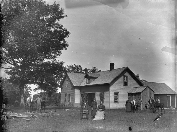 In the center foreground in a yard a girl is posing standing flanked by a man and woman sitting. On the left a man is standing with a horse. On the right a man and woman are standing with a horse, and another man is standing with a horse and foal.