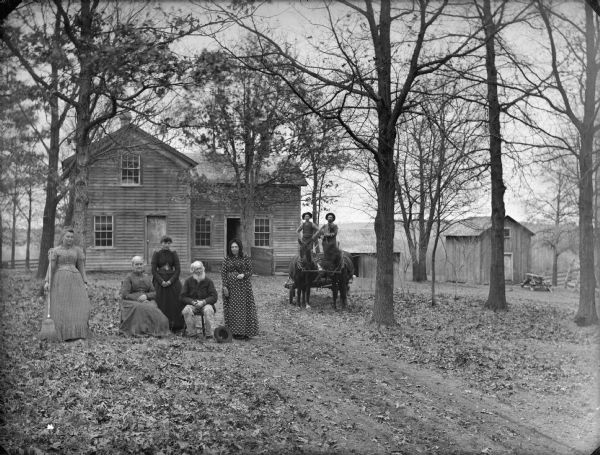 View down driveway towards a farm family posing in front of their farmhouse and farm buildings. An older man and woman are sitting. Three women are standing nearby, one of whom is holding a broom. Two men are standing on a horse-drawn wagon on the right.