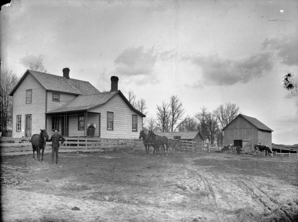 A man is displaying a horse near an empty buggy pulled by a team of two horses in front of a wooden fence. Just behind the fence is a frame house with a woman posing sitting on the porch. Cows are grazing in the yard on the right near farm buildings.