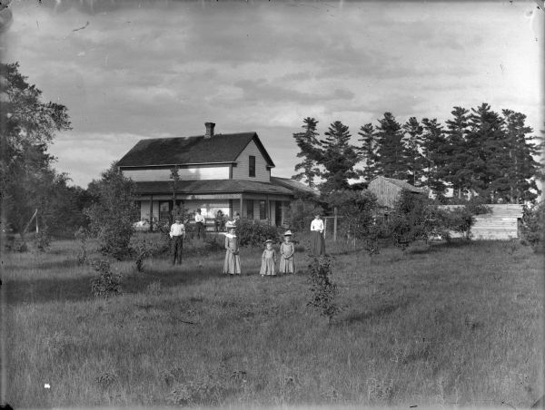Two men, three girls, and a woman are posing standing in a yard in front of a frame house farm buildings. In the background a man and woman are posing sitting on the porch.