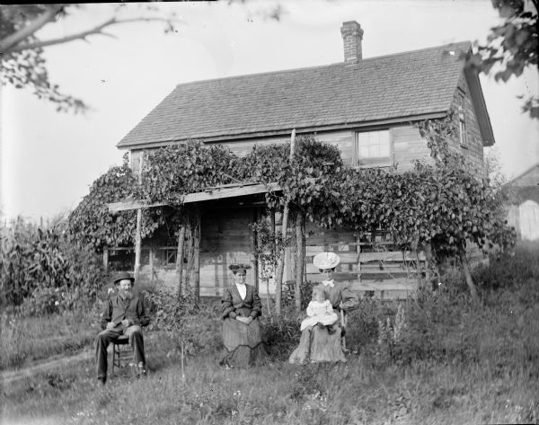 A man and two women, one holding a small child in her lap, are posing sitting in a yard in front of an arbor and frame house.