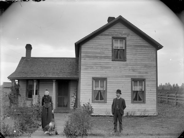 Man, woman, and dog posing standing in a yard and on the board sidewalk in front of a frame house.	