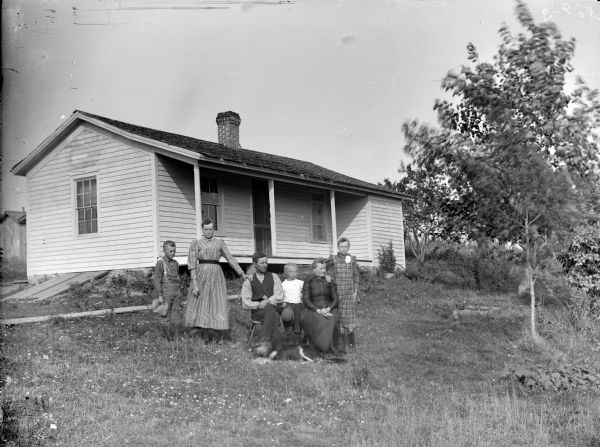 In the yard in front of a house a man and woman are posing sitting. Two girls and two boys are posing standing.