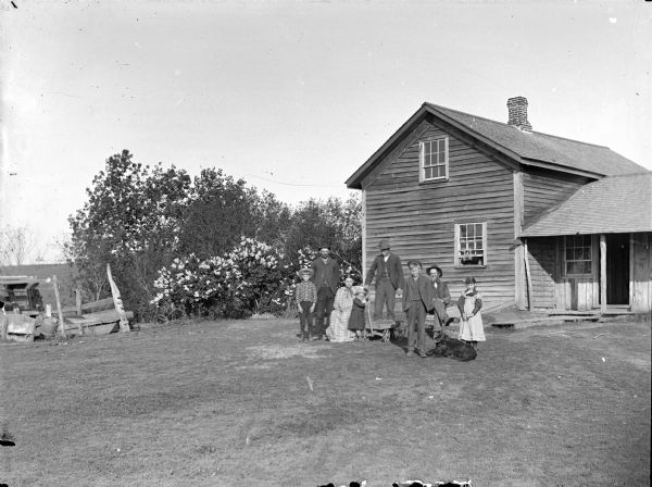 View across yard of elderly man and woman posing sitting, with two men, two boys, a girl and small child with a wagon posing standing. The group, along with a dog, is front of a frame house.