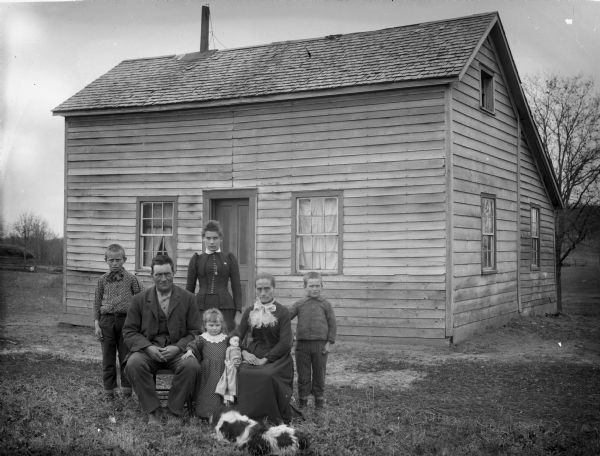 A man, woman, mother-in-law, and three children posing in the yard in front of a clapboard frame house. The young girl in the middle is holding a doll, and a spotted dog is standing in front of the group.