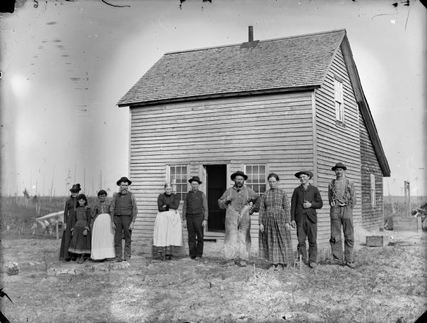 Five men, four women, and a girl are posing standing in front of a frame house and a well. The man on the far right is carrying a heavy chain over his shoulder.