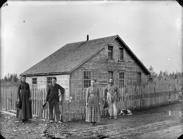 Two women, a man and a boy are posing standing next to a fence surrounding a frame house. On the right a dog is curled up at the base of the fence.