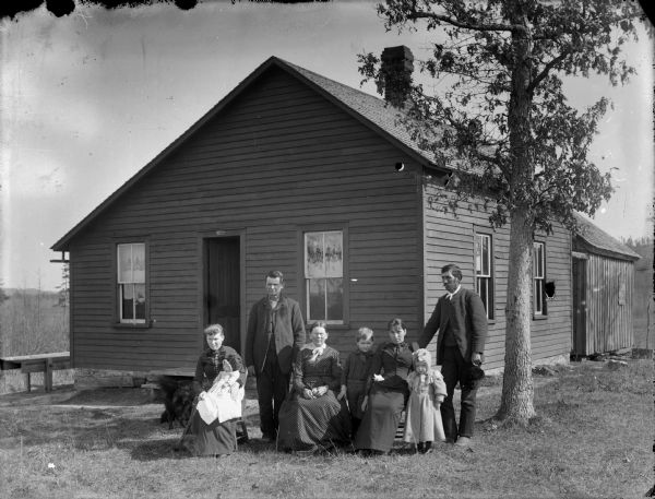 Group portrait of three women, one of them holding a baby, sitting in chairs, with two men, and a young boy and girl posing standing. The group is in a yard along with a dog in front of a frame house. Small sign above doorway reads: "Phoenix of Hartford."