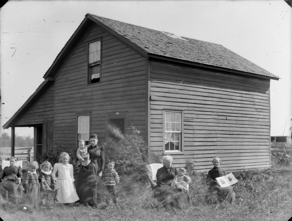 Three women, one holding a child, are posing sitting in chairs in the yard of a frame house, along with a woman holding an infant, three girls, and one boy standing. The woman sitting on the far right is holding open a photograph album. There is an insurance plaque above the doorway.	