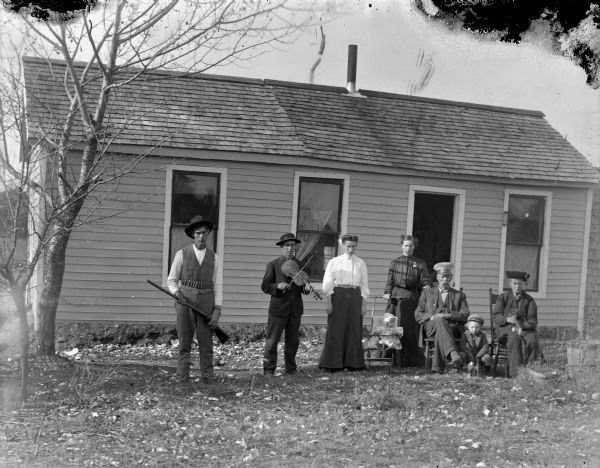 Eight people posing in front of a one-story clapboard house holding possessions. The man on the far left is holding a gun and wears a ammunition belt around his waist. The man next to him is holding a fiddle or violin. Two women in the middle are standing near an infant in a baby stroller. A man wearing a hat sits in a chair smoking a pipe. On the far right an older woman wearing a dark dress and a hat is sitting in a chair holding a puppy. Next to her a small boy sitting on the ground is also holding a puppy.