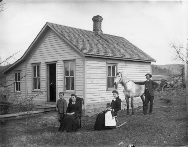 Group posing in front of house. On the right, a man is standing with a white horse, and behind him is a carriage. Two women are sitting in rocking chairs, with a young girl and boy standing in the yard.