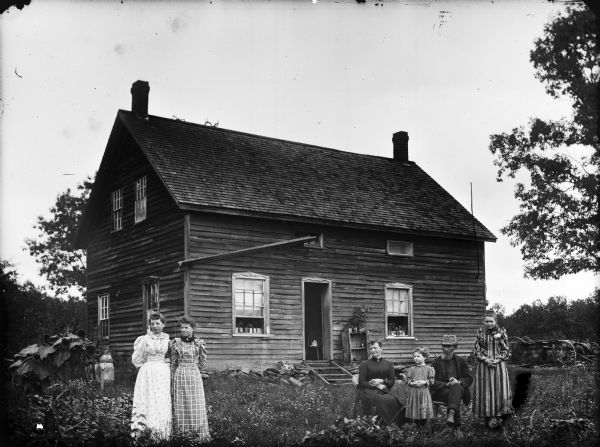 Man and woman posing sitting in a yard in front of a two-story frame house, with a young girl and a young woman standing next to them. On the far left two other young women are standing together. They are probably Bob Farrington and family. From left to right: Azalea Mead, Elsie Mead, Mrs. Farrington, Edna Farrington, Bob Farrington, and unknown.	