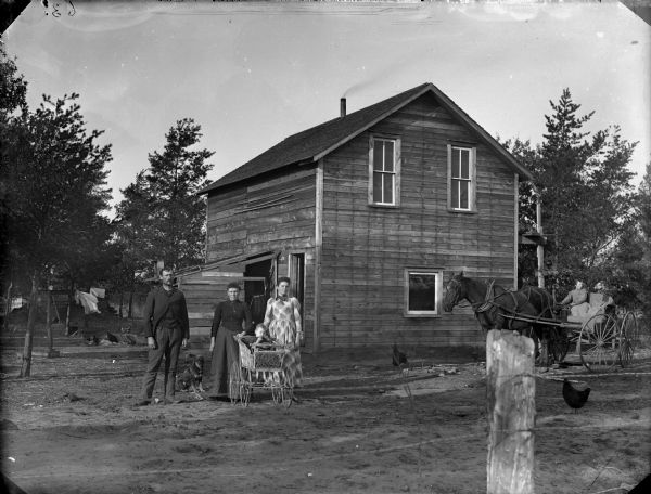 Two women, a man, and a dog are standing next to a baby who is sitting in a baby carriage. The group is in front of a two-story frame house. On the far right a boy is posing sitting in a buggy pulled by a single horse. There is a wire fence in the foreground, and chickens are wandering about the yard.	