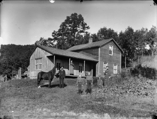 Group of people standing posing in a yard, and one woman in the background standing on the front porch of a frame house which is on the slope of a hill. In the yard on the left is a man displaying a horse, in the center are two boys, and on the right is a woman and a young girl.
