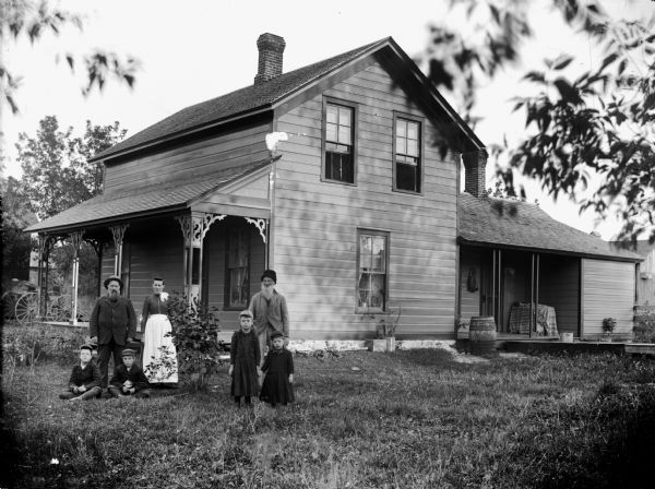 Group of people posing in front of frame house. Two men, two girls, and a woman are standing, and two boys are sitting on the ground. They are all wearing hats except the woman, who is wearing a white apron and has a scarf tied around her neck. There is a carriage parked in the background on the left at the side of the porch which has eave woodwork decoration. On the right is a barrel near another covered porch area with wooden walkway.
