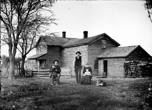 A family of five posing with their dog in the yard in front of their house. On the left is an older man sitting in a chair with a child in his lap, and in the center a younger man is standing with his hand on a chair where a woman is sitting with an infant on her lap. Behind them is a wood frame house surrounded by a fence, with an attached outbuilding, and another barn or shed in the far background.