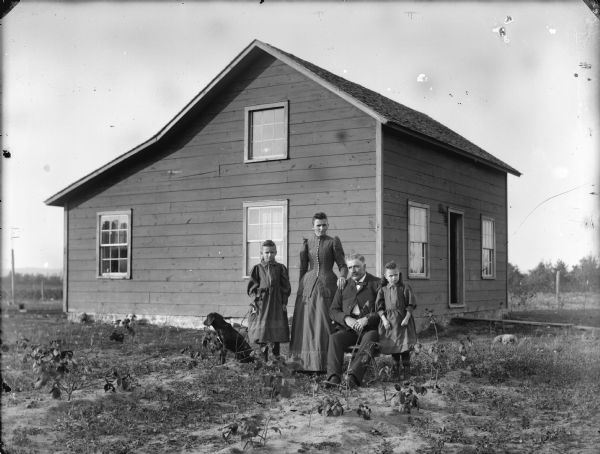 Group of people posing in yard in front of a small frame house. In the center is a man sitting, with two young girls and a woman standing beside him. A dog is standing to the left of the group. Grass and small plants are scattered in the yard, and small trees are in the far background.