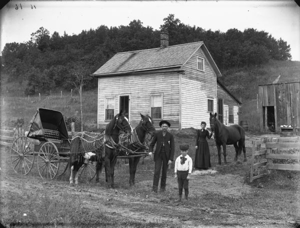 Three people are standing in front of a frame house which is at the foot of a small tree-covered hill. On the left a man is standing with a team of two horses wearing fly-nets who are hitched to an empty buggy. On the right a woman is standing displaying a horse, and in the center is a small boy standing. In the background on the right is a small barn or shed, with a hand-pump nearby.