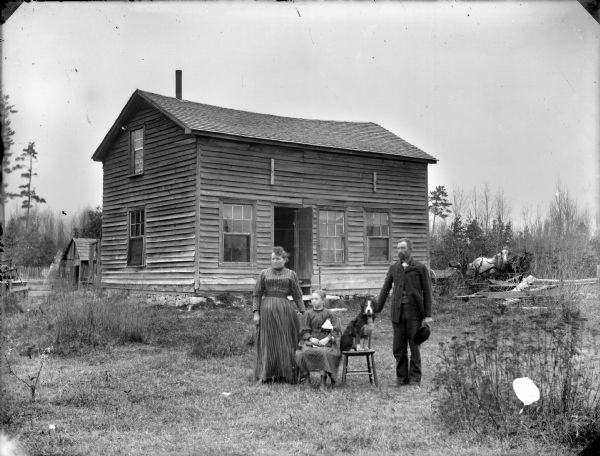Group posing in the yard front of a frame house. A woman is standing next to a girl who is sitting and holding a doll. In the center is a dog sitting on a chair. Next to the dog on the right is a man standing holding on to the back of the chair. In the background on the right behind the house is a team of two horses hitched to a wagon.