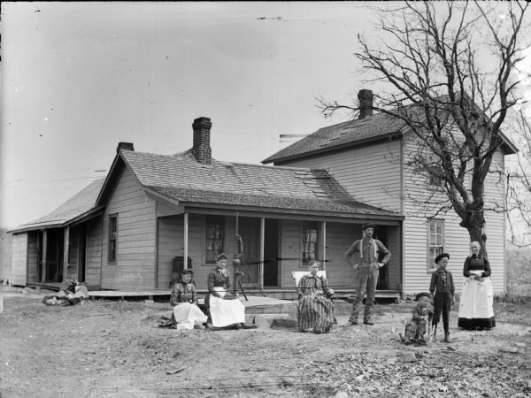 Group portrait of people posing in the yard in front of a frame house, with a porch and a hand-pump. There are three women sitting on the left, and a small boy sitting in a small rocking chair on the right. Standing on the right behind the small boy are a woman, man, and a young boy. There is a dog sitting in the open doorway on the porch. Everyone but the man is either holding flowers or are wearing them on their lapels.