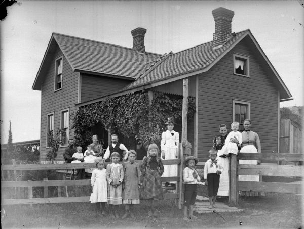 Group portrait of people posing in front of a frame house with a porch covered in vines. In the foreground four young girls are in front of a fence, and two young boys are standing near the gate. Standing next to the boys are two women, with one woman holding a young child perched on the gatepost. Another woman is standing on the wooden walkway leading to the porch. Behind the fence in the yard near the porch are two men and a woman sitting, two of them holding two small children in their laps.
