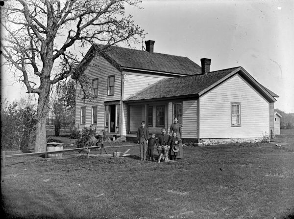 View of group of people posing in a yard. A man is sitting surrounded by a woman, young girl and boy, and an older boy. The young girl is holding a hat and has a doll cradled in her left arm. A dog is sitting in front of the man. Behind them is a frame house with a cat sitting on the porch. In the yard on the left is a water pump, two pails and a barrel.